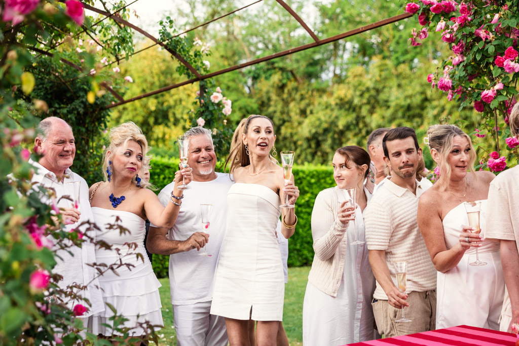 Group of people in white clothing holding champagne glasses at a garden party with pink flowers, evoking the elegance of a Tuscany wedding.