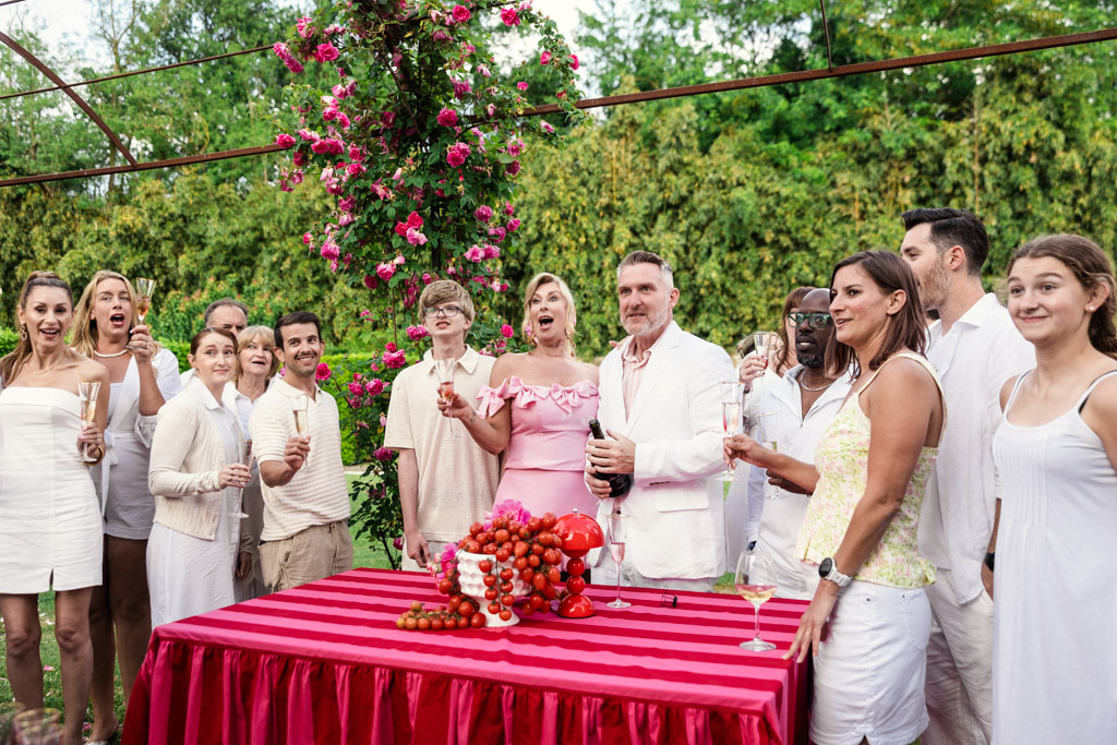 Group of people in white outfits, celebrating outdoors around a red table with champagne and grapes, capturing the joyful spirit of a Tuscany wedding.