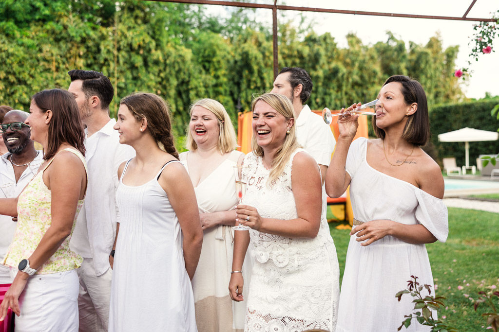A group of people wearing white clothes laugh and drink outside at a garden party, evoking the joyful and elegant atmosphere of a Tuscany wedding.
