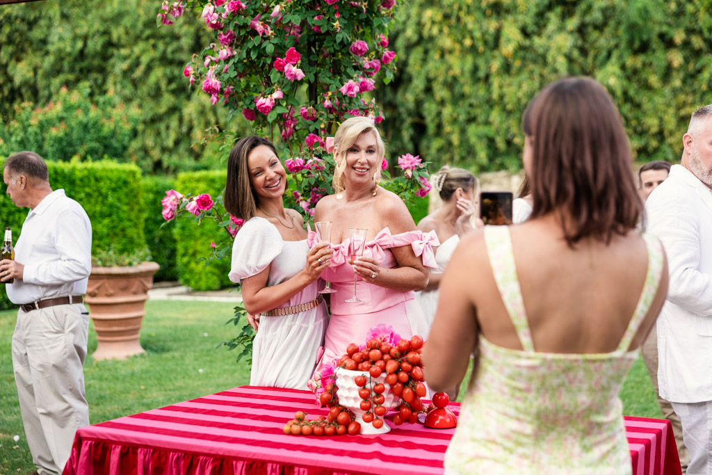 Two women pose and smile for a photo at an outdoor party by a table with flowers and tomatoes, capturing the joyful spirit of a Tuscany wedding.