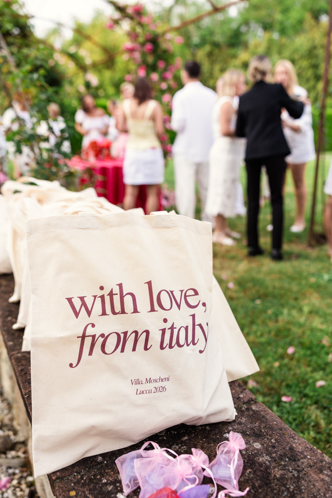 Canvas tote bag reading “with love, from italy” at an outdoor gathering, reminiscent of a Tuscany wedding, with people in white in the background.