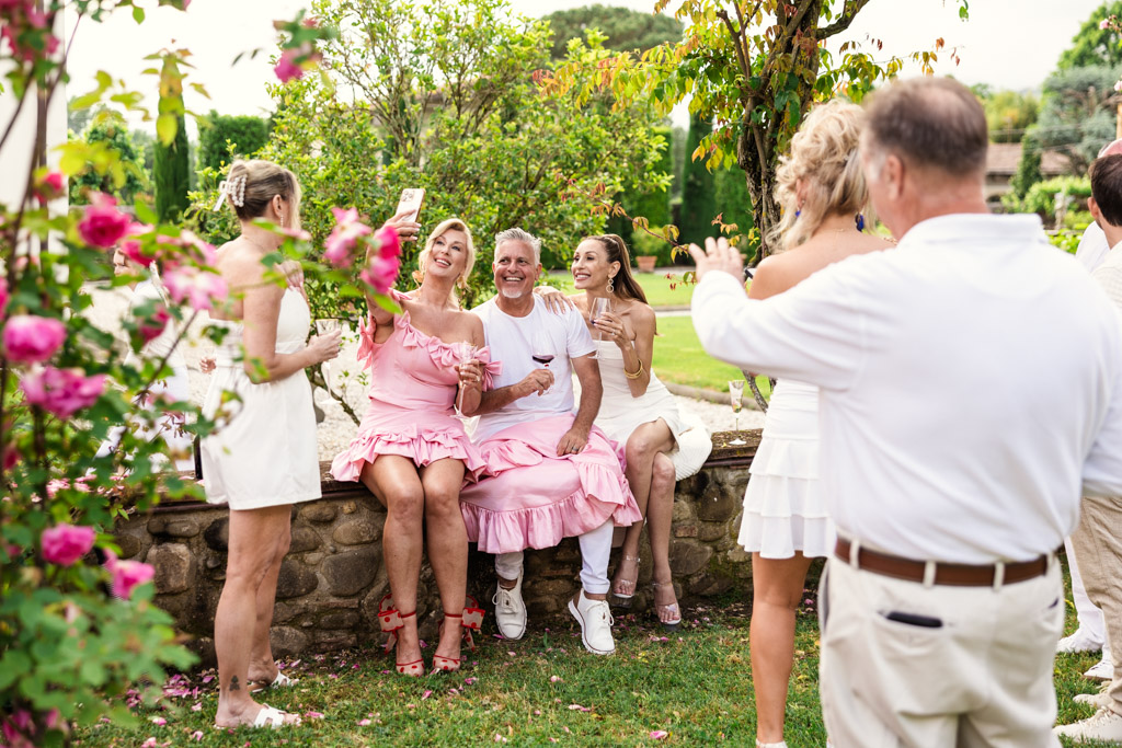 People in pink and white outfits smile, pose, and take photos in a garden at an outdoor gathering reminiscent of a Tuscany wedding.