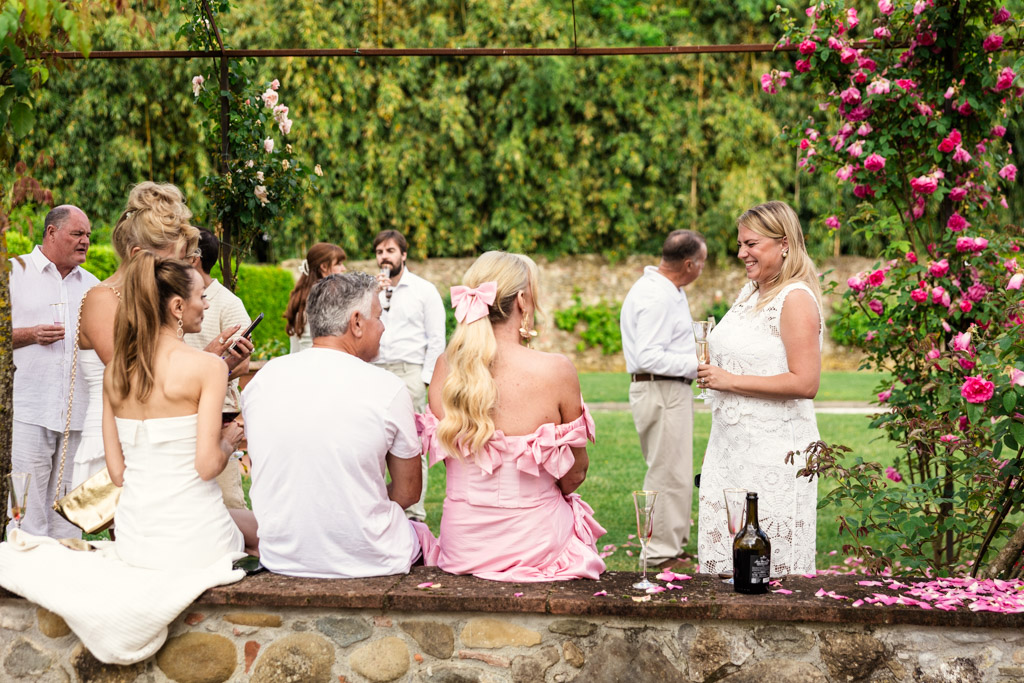 People in white and pink outfits converse outdoors by roses, holding drinks and enjoying a sunny garden gathering reminiscent of a charming Tuscany wedding.