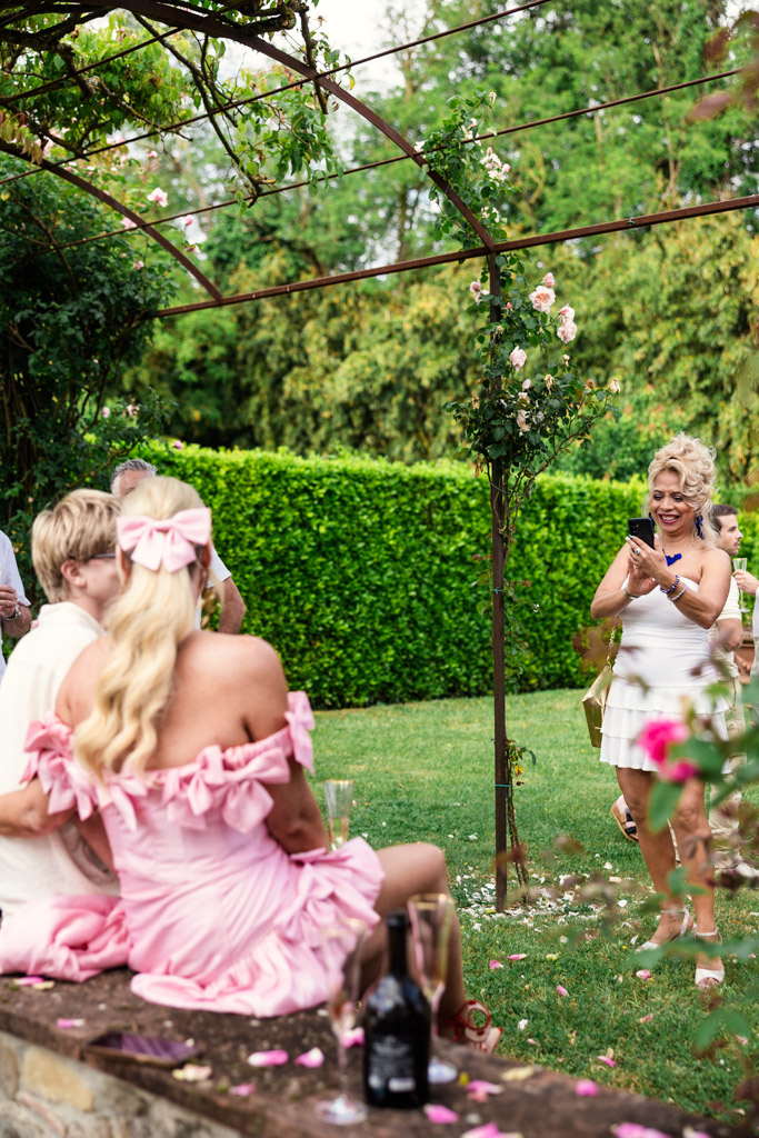 Woman in white dress takes photo of two people in pink dresses sitting on a garden wall at an outdoor Tuscany wedding.
