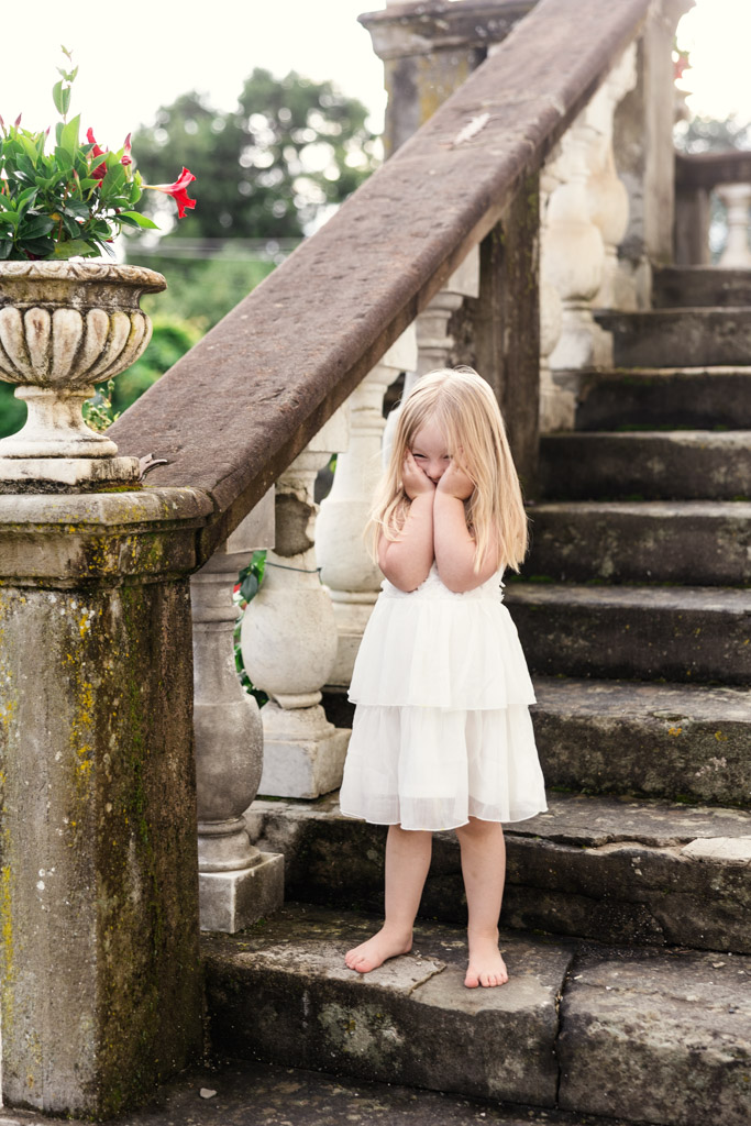 Barefoot girl in a white dress stands by old stone stairs, covering her face with her hands, outdoors—a scene reminiscent of a quiet moment before a Tuscany wedding.