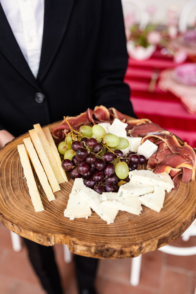 Person holding a wooden platter with cheese slices, prosciutto, and green and red grapes—a delightful spread reminiscent of a Tuscany wedding.