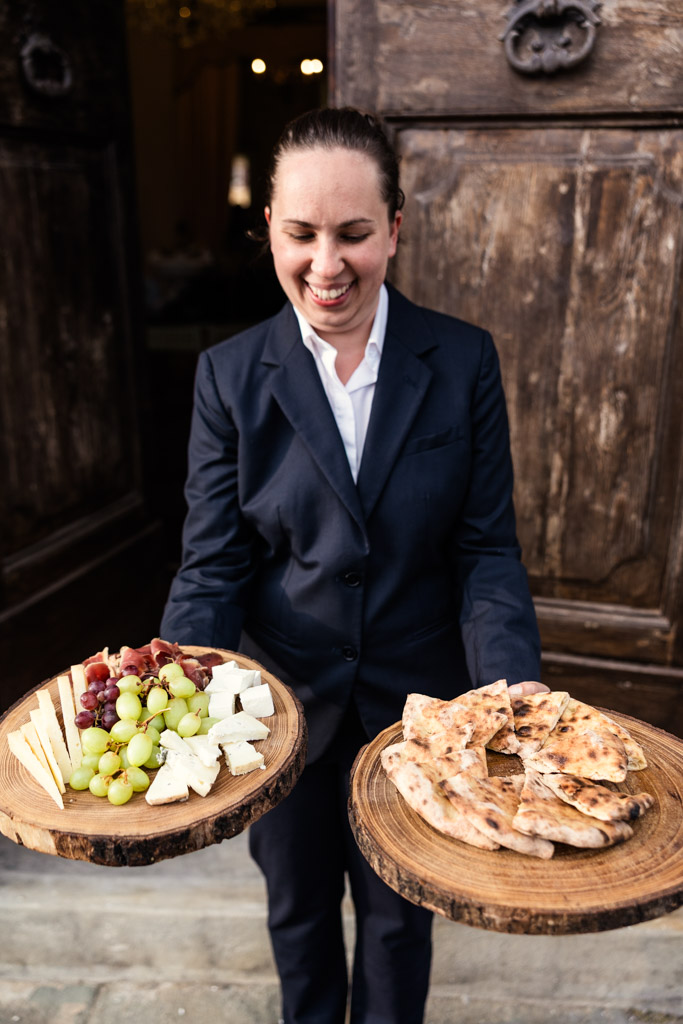 A smiling server in a suit holds wooden trays with cheese, grapes, and sliced bread in front of rustic doors, capturing the warm hospitality of a classic Tuscany wedding.