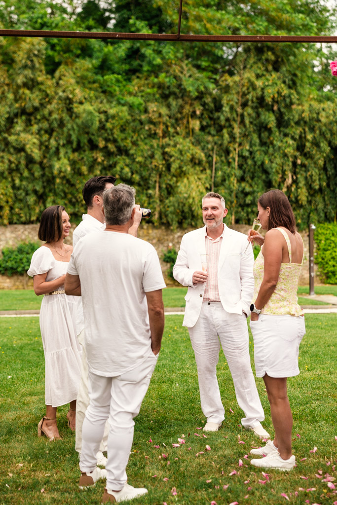 Five adults in white attire stand on grass chatting, with lush greenery and trees in the background, evoking the elegance of a Tuscany wedding.