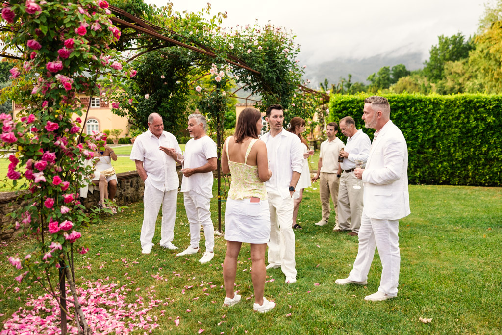 People in white outfits gather and talk in a garden near pink flowers and greenery on a cloudy day, evoking the charm of a Tuscany wedding.