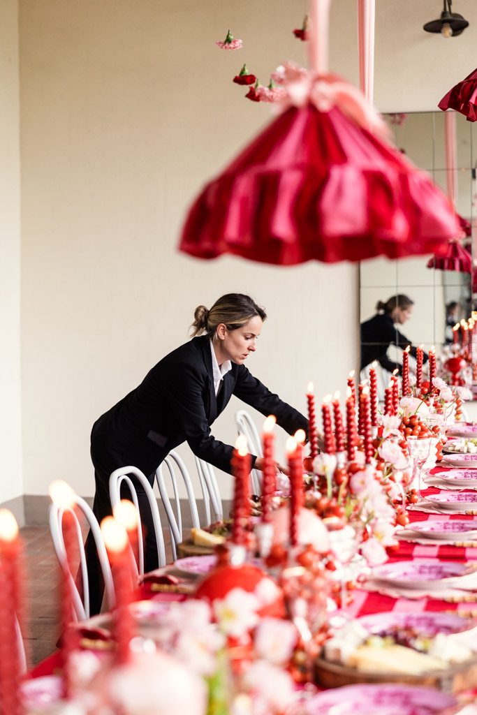 A woman arranges a long table set with red candles, pink flowers, and red hanging decorations for a romantic Tuscany wedding.