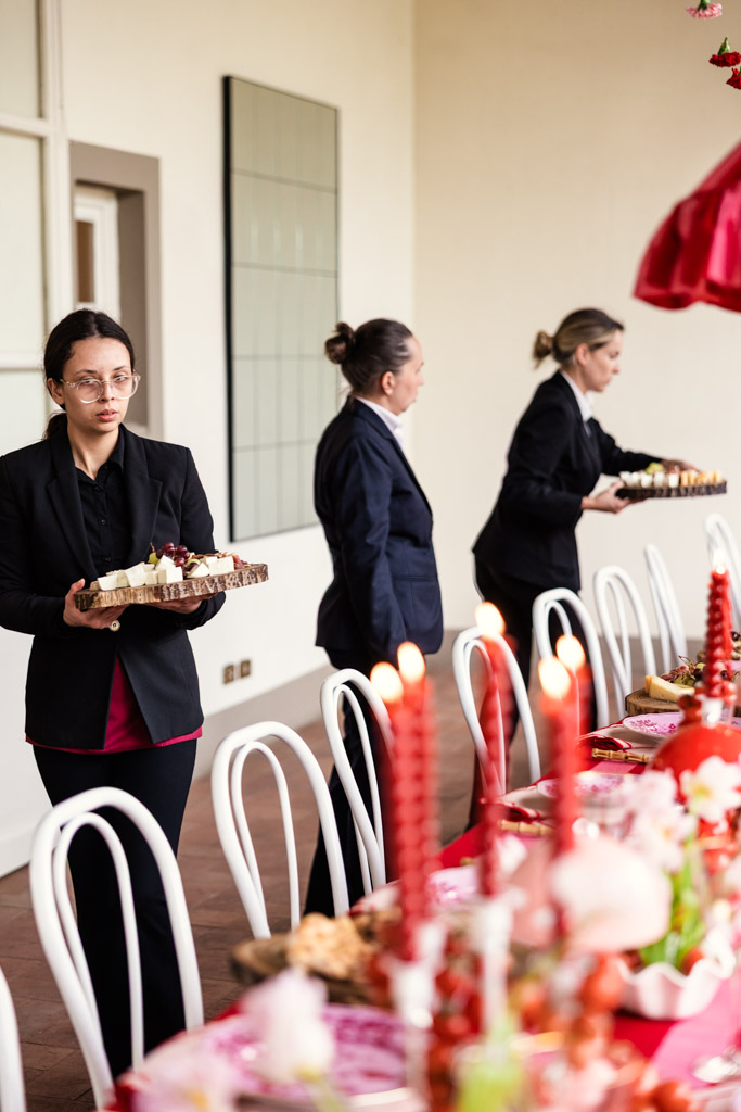 Three women in black suits serve food at a long, festively decorated table with red candles, capturing the elegant atmosphere of a Tuscany wedding.
