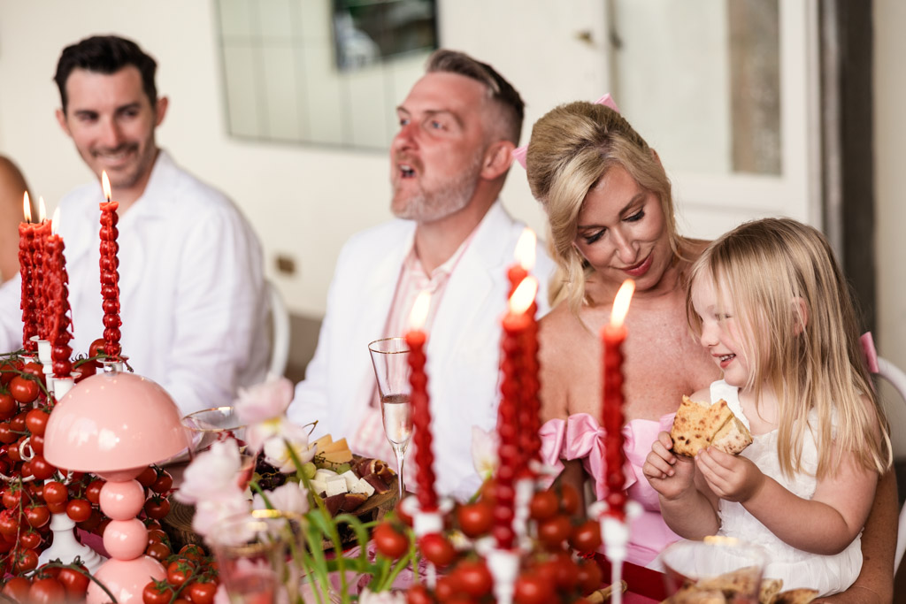 A family sits at a festive table with red candles, eating and smiling together, reminiscent of the warmth and joy found at a Tuscany wedding.