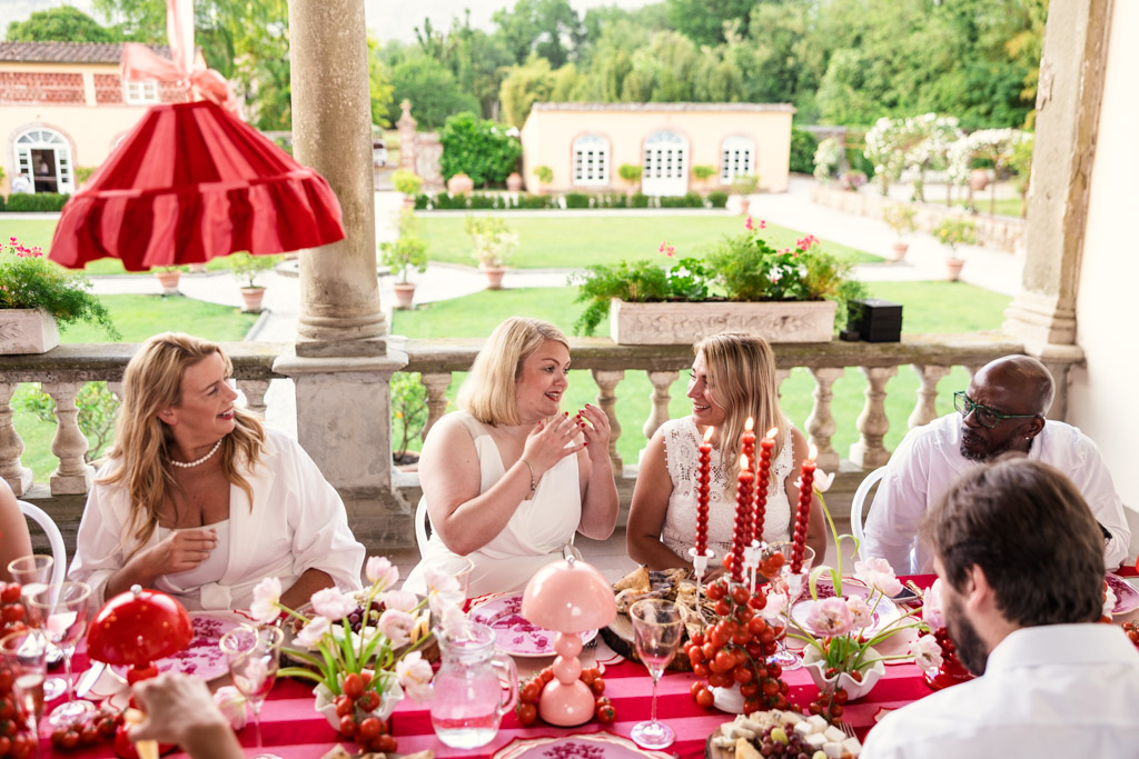 People in white clothes sit at a festive outdoor table, talking and laughing, with red and pink decorations reminiscent of a joyful Tuscany wedding.
