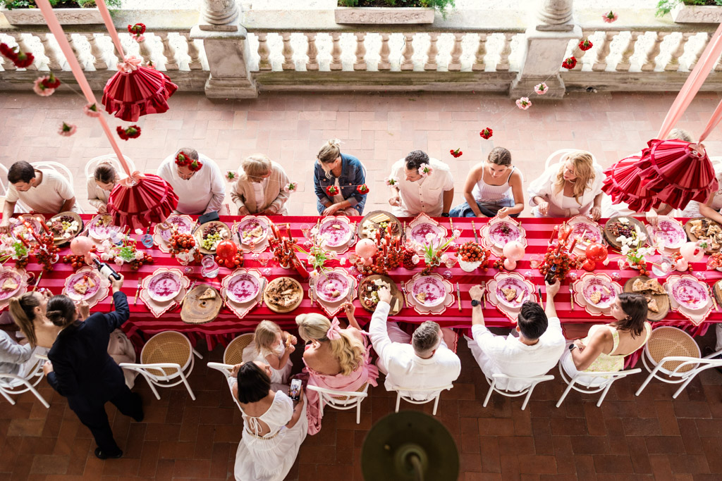 People dining at a long, festive table with pink decorations and food, viewed from above, capturing the joyful atmosphere of a Tuscany wedding.