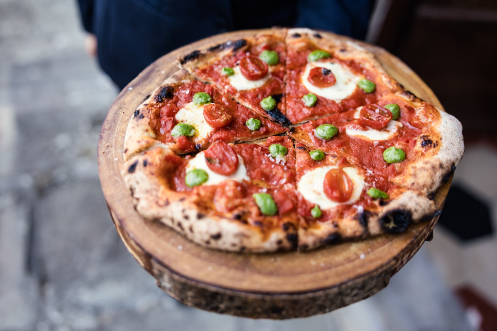 A wood-fired pizza with tomatoes, cheese, and green toppings on a wooden platter—perfect for sharing at a cozy Tuscany wedding.