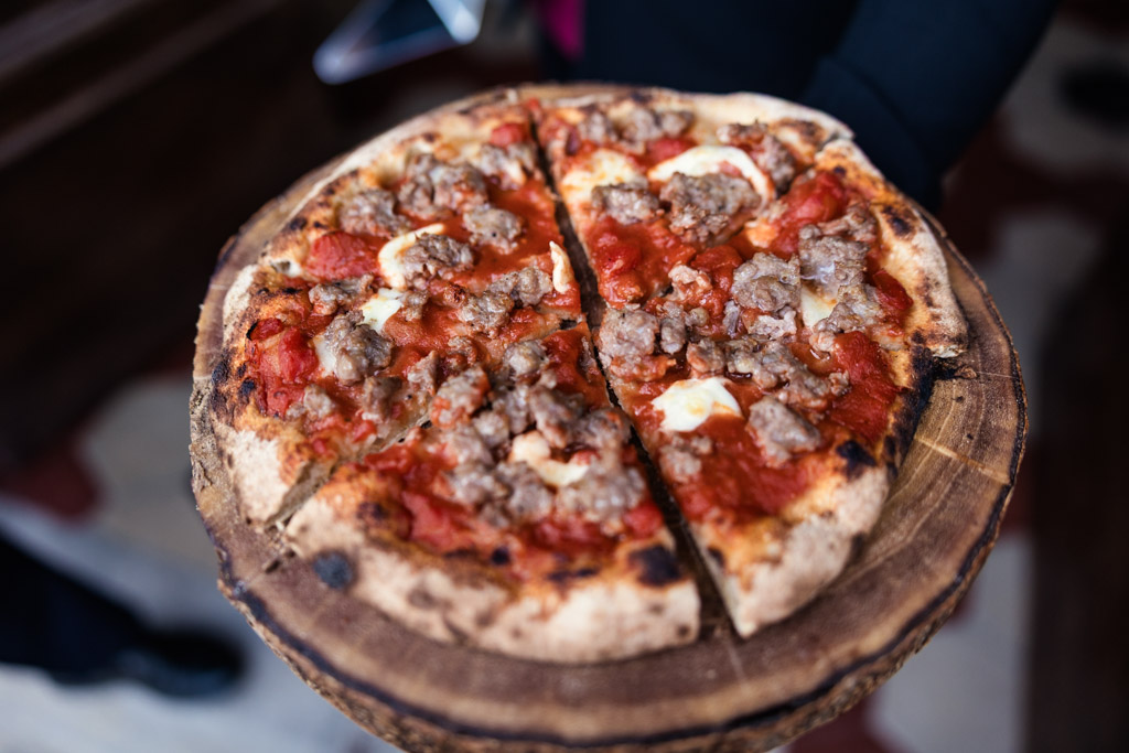 A close-up of a sausage pizza cut into four slices, served on a rustic wooden platter—perfect for sharing at an intimate Tuscany wedding.