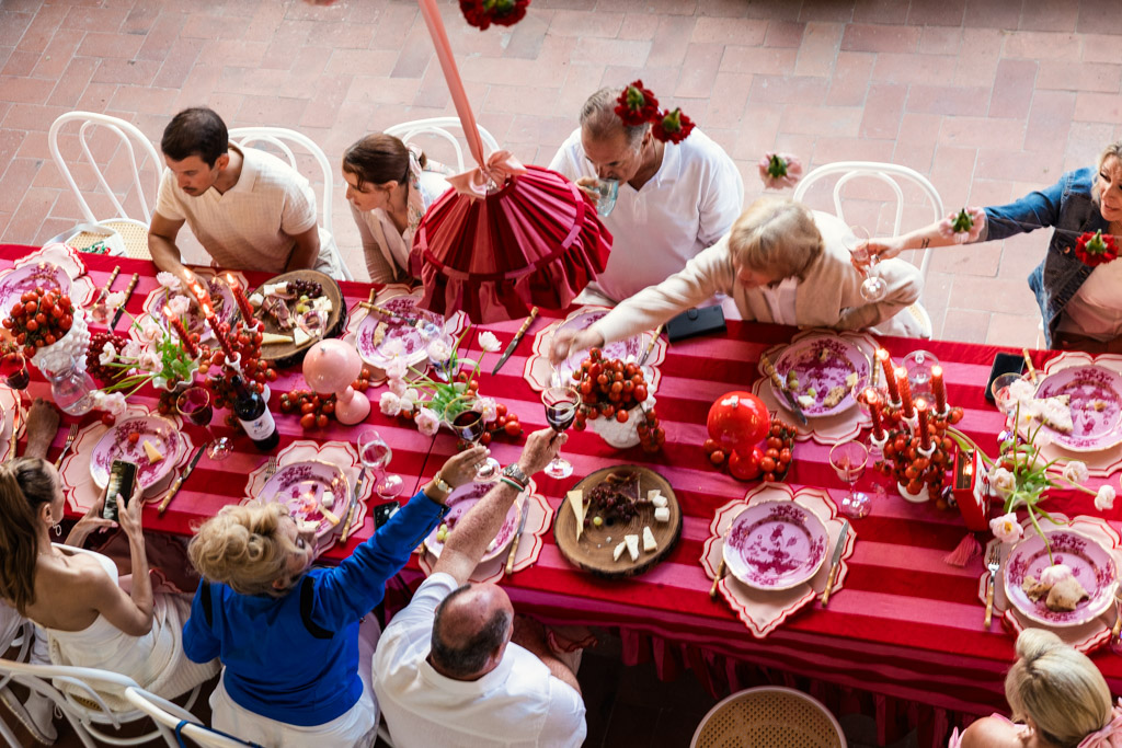 People sharing food and drinks around a red-striped table set for a festive meal, seen from above—a scene reminiscent of a joyful Tuscany wedding.