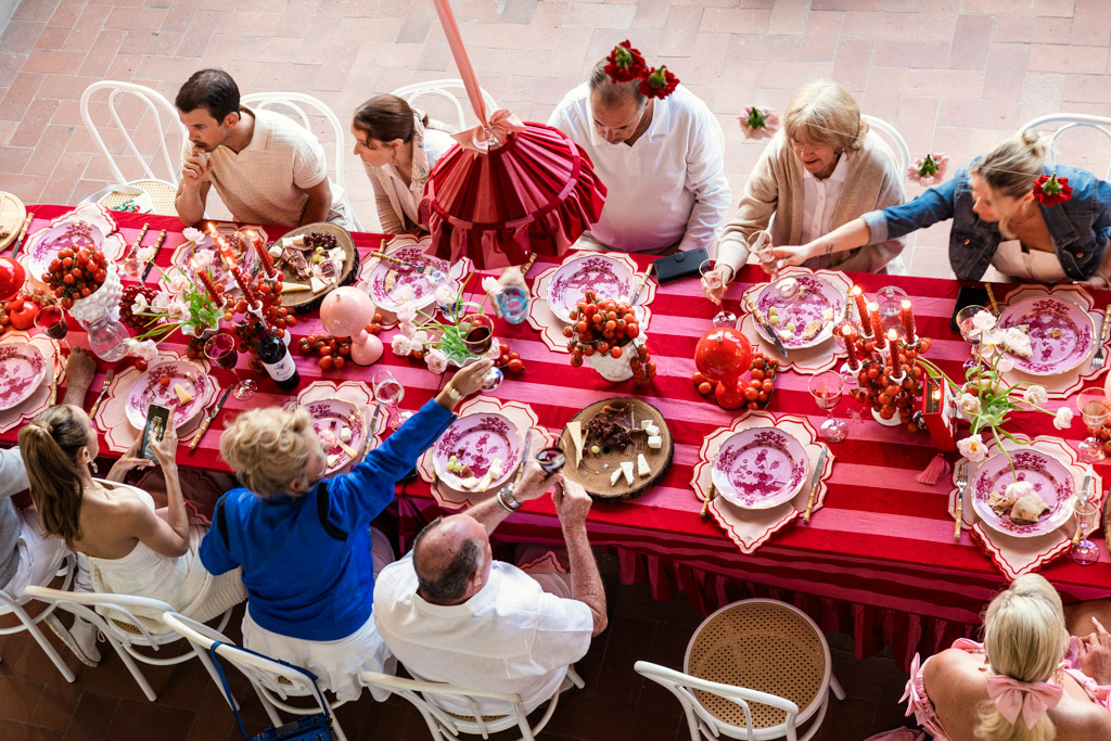 People gathered around a long red table, sharing food and taking photos at a festive meal reminiscent of a Tuscany wedding.