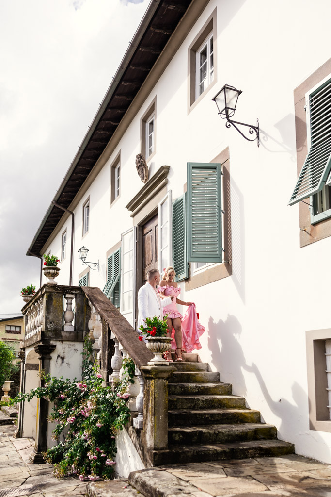 A couple in pink and white sits on stone steps outside a white house with green shutters and potted flowers, capturing the romantic charm of a Tuscany wedding.