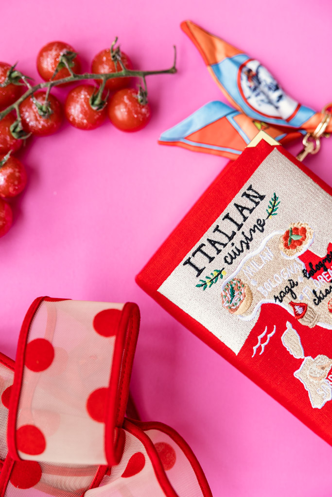 Red Italian cuisine notebook, polka dot ribbon, tomatoes, and a colorful scarf evoke the charm of a Tuscany wedding on a pink background.