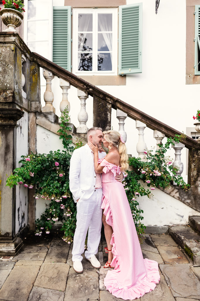 A couple shares a kiss on a stone patio near stairs, the woman in a pink dress and the man in white, evoking the romance of a Tuscany wedding.