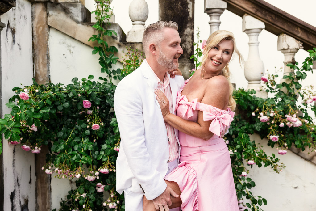 Smiling couple in pastel pink outfits posing by a stone staircase with blooming roses in the background, capturing the romantic charm of a Tuscany wedding.