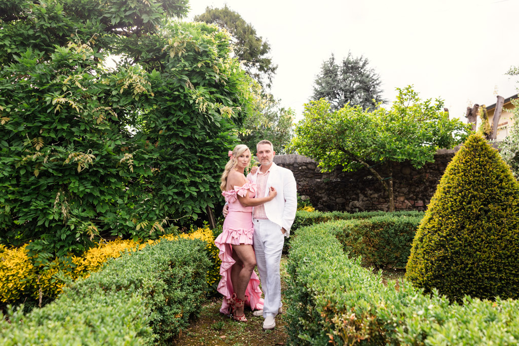 A woman in a pink dress and a man in white stand together in a lush, manicured garden, evoking the romantic atmosphere of a Tuscany wedding.