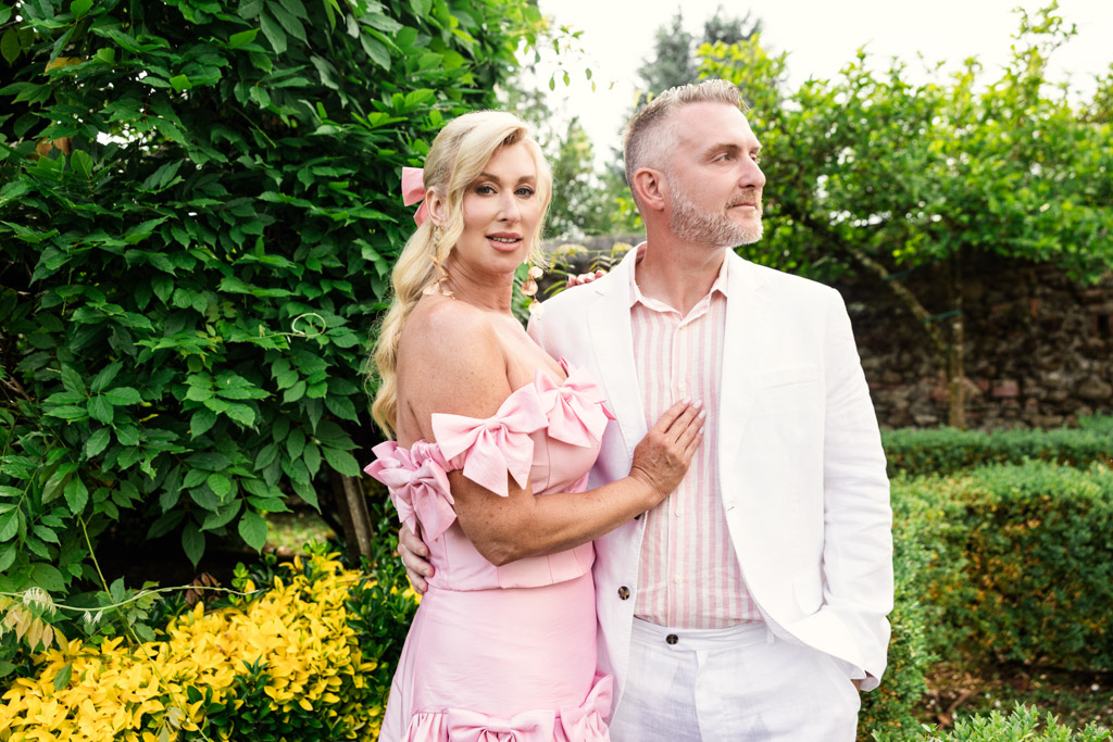 A woman in a pink dress and a man in a white suit pose together in a lush garden, capturing the elegance and romance of a Tuscany wedding.