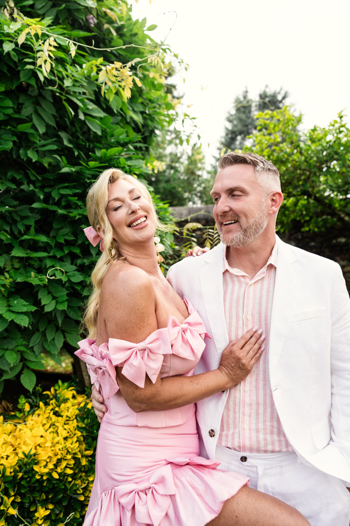 Smiling couple poses outdoors at their Tuscany wedding, woman in pink dress, man in white suit, surrounded by green and yellow plants.
