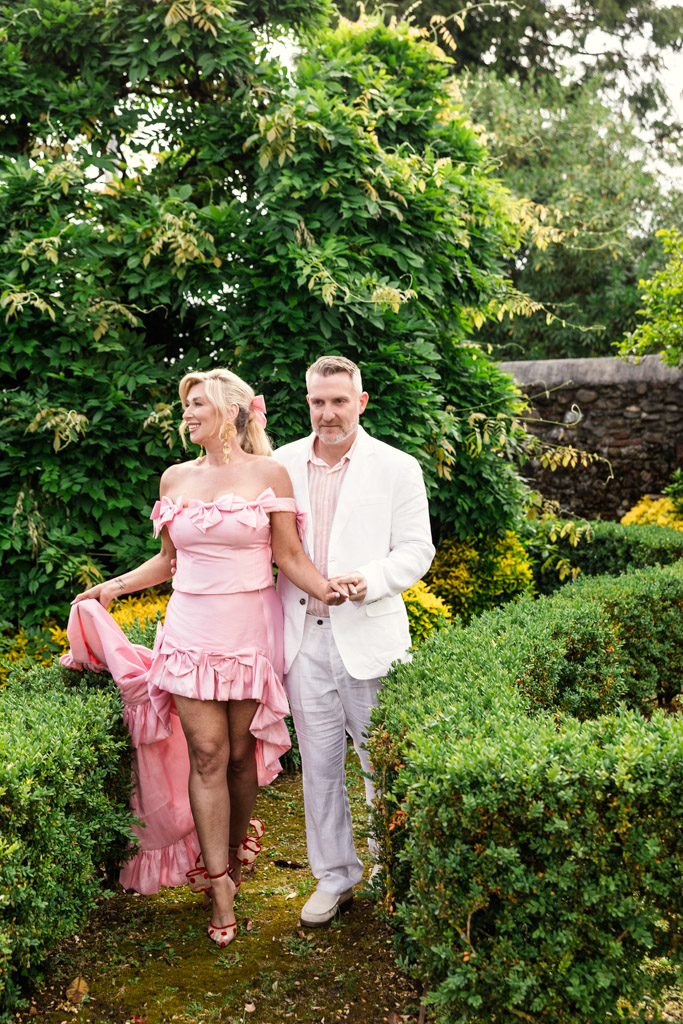 A woman in a pink dress and a man in a white suit walk together in a lush green garden, capturing the romance of a Tuscany wedding.