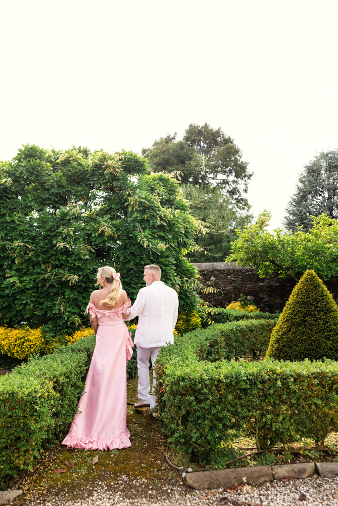 A couple walks hand in hand through a manicured garden, dressed in light pink and white formal attire, evoking the effortless romance of a Tuscany wedding.