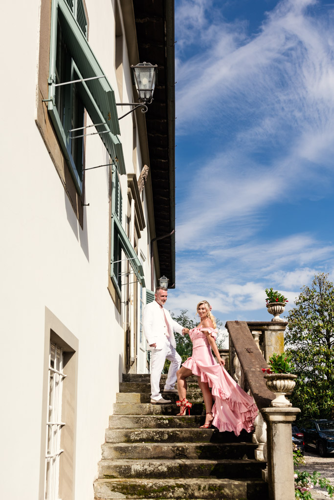 A man in white and a woman in a pink dress stand on sunny stone steps beside a white building, capturing the romance of a Tuscany wedding.