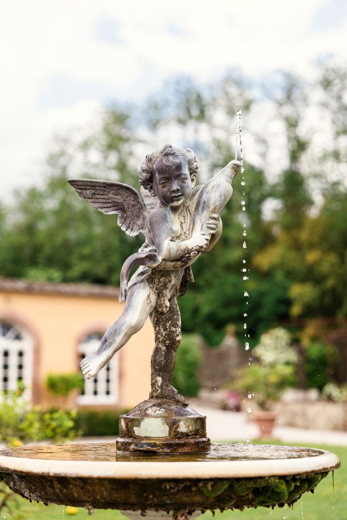 An angel statue holding a fish spouts water in a garden fountain, with blurred greenery and a building behind, creating an enchanting scene reminiscent of a Tuscany wedding.