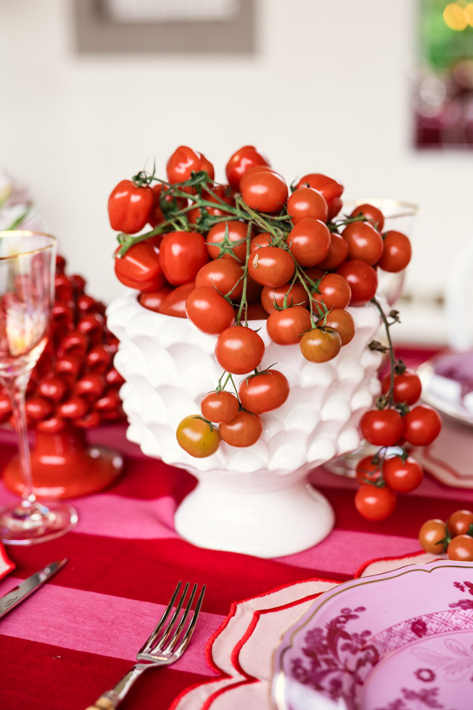 Cherry tomatoes piled in a white bowl on a red and pink tablecloth with plates and glassware nearby, evoking the vibrant charm of a Tuscany wedding tablescape.