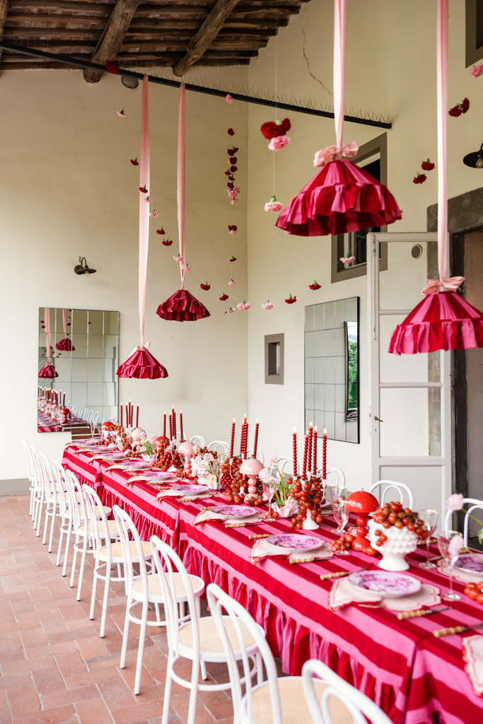 A long table with red and pink decor, hanging ribbons, candles, and white chairs set for a festive meal evokes the charm of a Tuscany wedding.