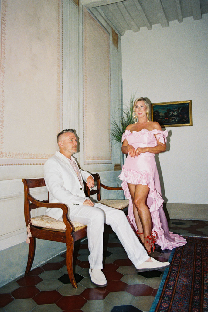 A man in a white suit sits on a bench while a woman in a pink dress, evoking the elegance of a Tuscany wedding, stands beside him indoors.