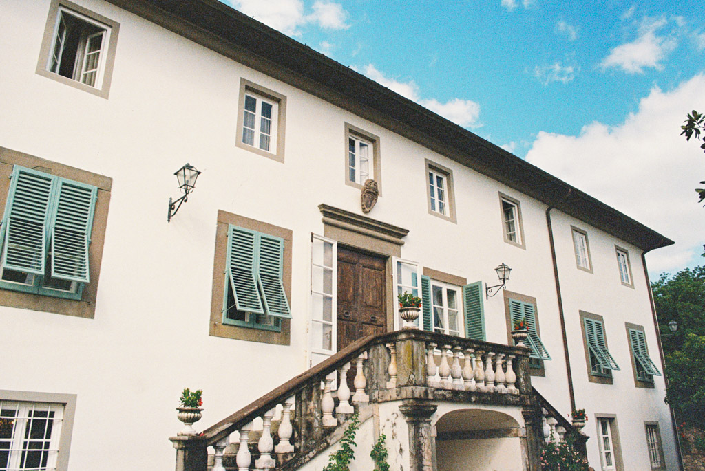 Large white house with green shutters, stone stairs, and potted plants under a partly cloudy sky, perfect for a romantic Tuscany wedding.
