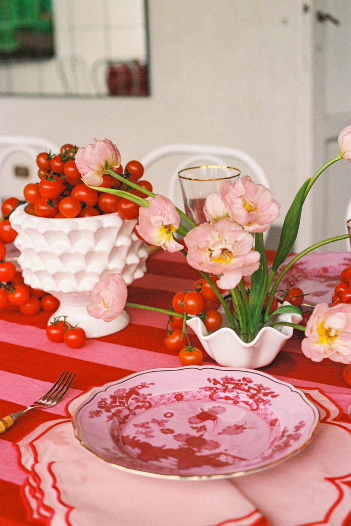 Pink tulips and cherry tomatoes in white vases on a red striped tablecloth with a pink floral plate, evoking the rustic charm of a Tuscany wedding.