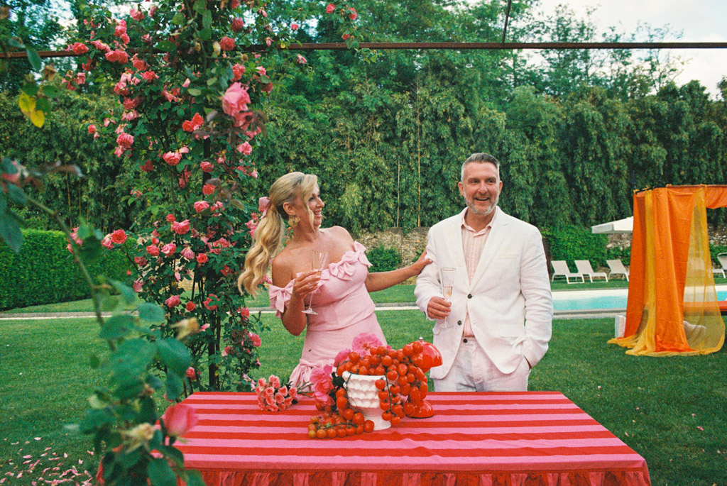 A smiling couple in pink outfits stands by a striped table with fruit in a lush garden setting, capturing the elegance and charm of a Tuscany wedding.