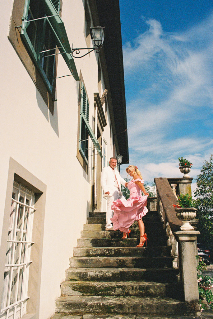 A man in white and a woman in a pink dress stand on stone steps outside a building under a blue sky, capturing the elegance of a Tuscany wedding.