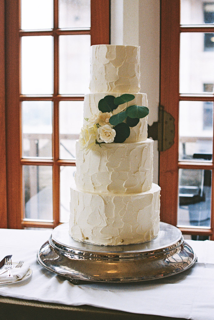 Film photo of four-tier white wedding cake with textured icing, white roses, and green leaves on a silver stand for wedding celebration at the University Club of Chicago