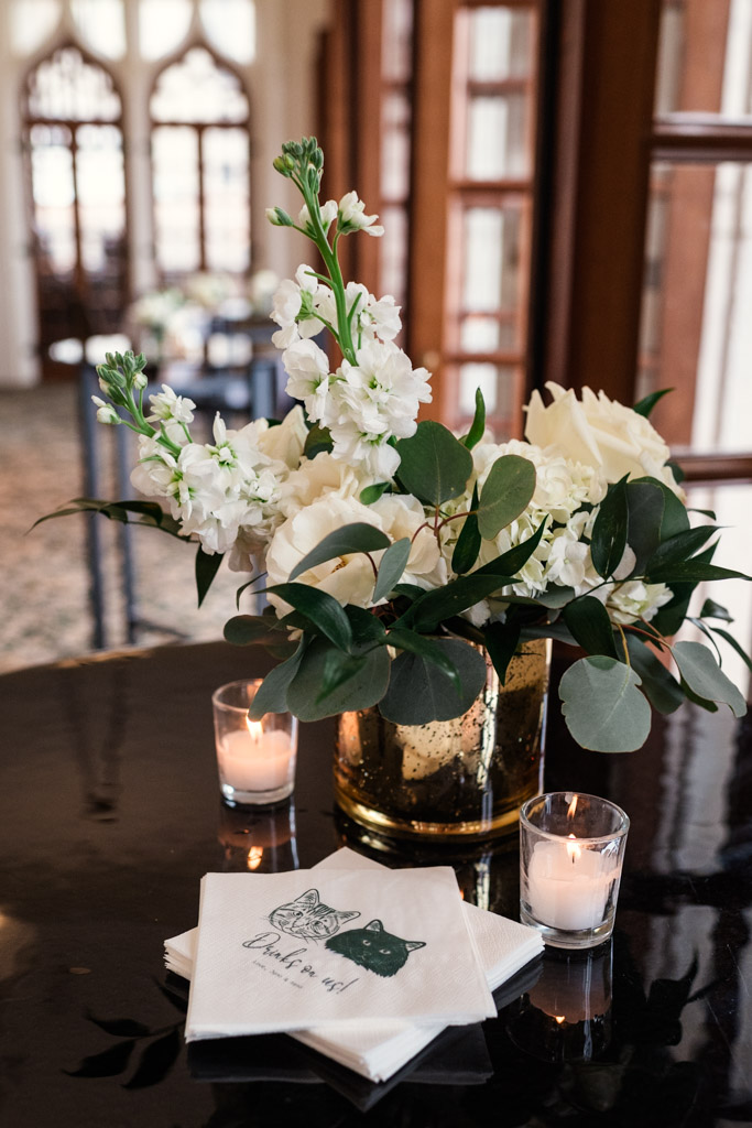 White flowers in a gold vase with candles and custom napkins adorn a dark table in a bright room for wedding celebration at the University Club of Chicago