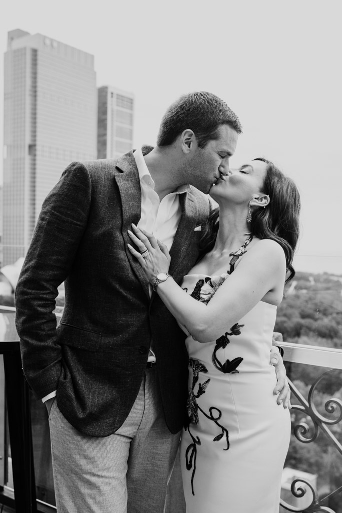 Black and white photo of newlywed couple kissing on a balcony at the University Club of Chicago, with city buildings in the background, both dressed in formal attire