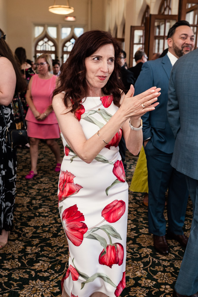 Woman in a white dress with red flowers clapping during wedding celebration at the University Club of Chicago, with people mingling in the background