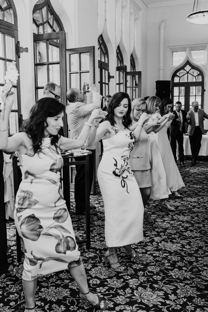 Black and white photo of bride and guests in formal attire dancing in a line at an indoor event at the University Club of Chicago, holding hands and smiling