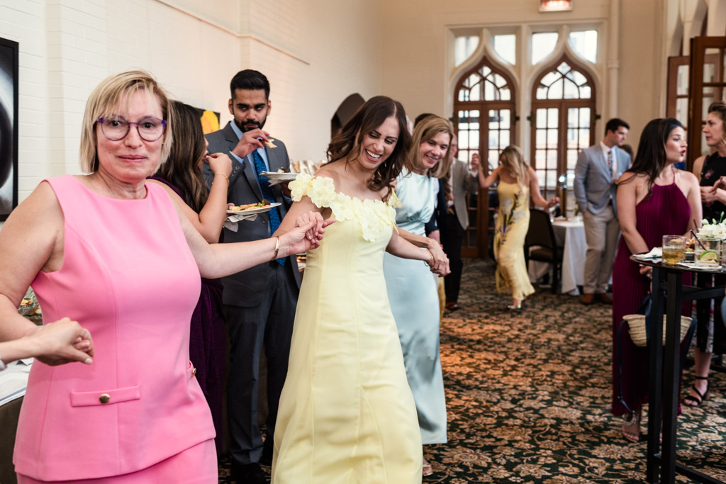 Guests in formal attire, holding hands and dancing during wedding celebration with arched windows in the background at the University Club of Chicago