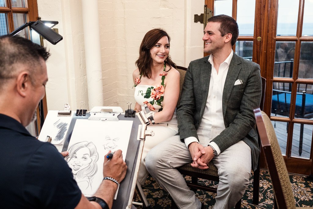 Newlywed couple sits smiling as an artist draws their caricature during their wedding celebration at the University Club of Chicago