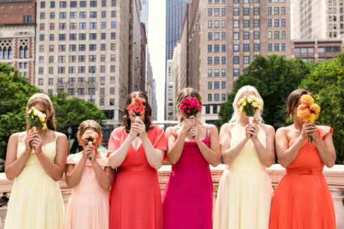 Six women in colorful dresses hold flowers in front of their faces near the Chicago Athletic Association.