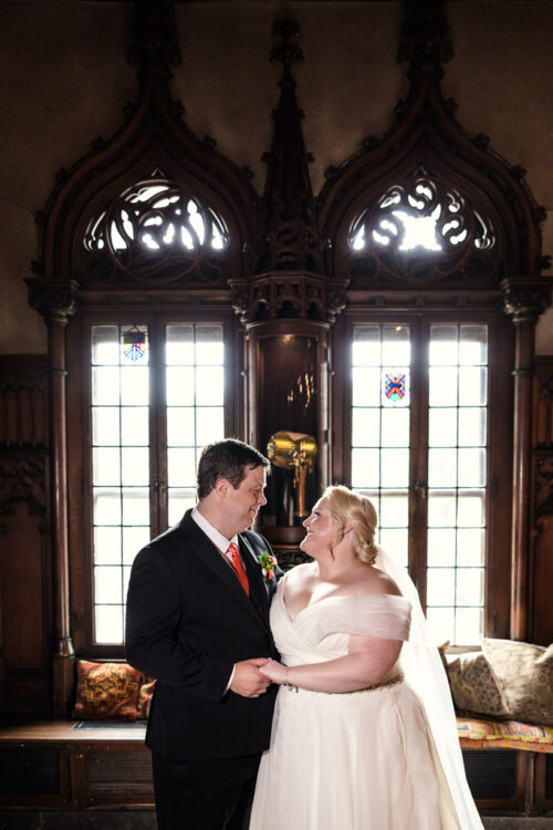 Bride and groom smiling at each other, standing before ornate windows at the Chicago Athletic Association.