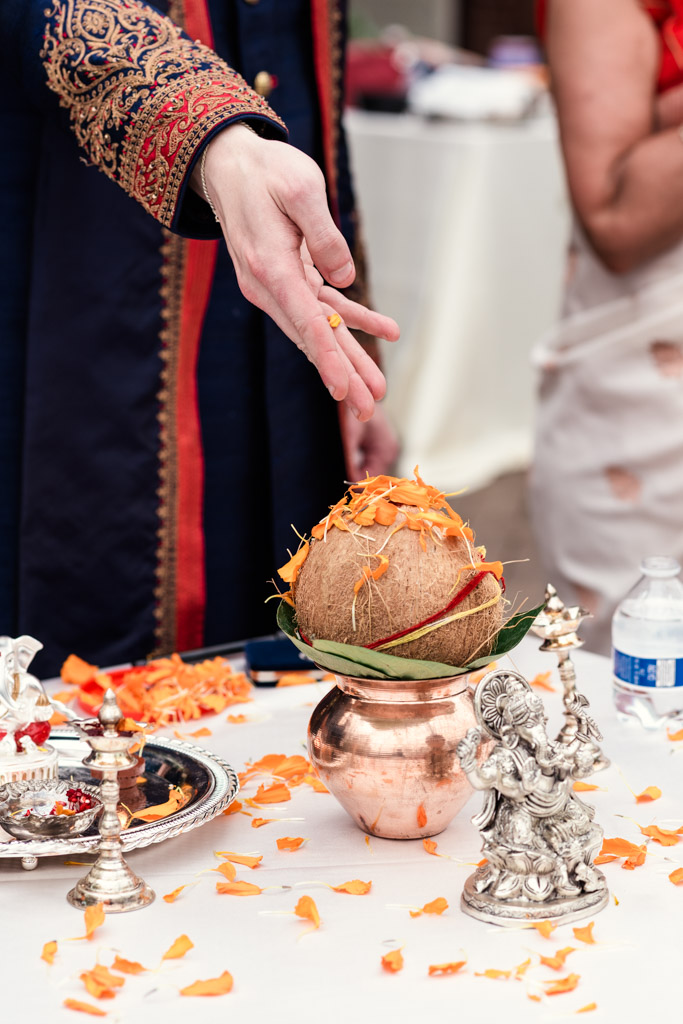 A hand sprinkles flower petals over a coconut in a copper pot next to a Ganesh statue during a traditional Chicago Indian wedding ritual.